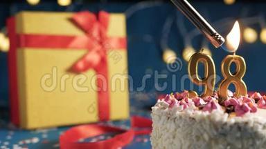 白色生日蛋糕数字98金蜡烛用打火机燃烧，蓝色背景灯和礼品黄色盒子用红色系起来