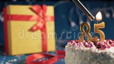 白色生日蛋糕，85根金色蜡烛，用打火机点燃，蓝色背景，黄色礼品盒装有红色