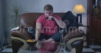 白人成年人看电视笑的肖像。 坐在沙发上吃<strong>薯片</strong>喝啤酒的单身男人