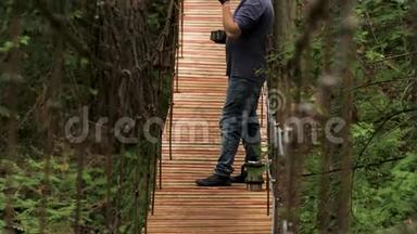 一个年轻的小伙子站在山林峡上的挂木桥上，<strong>喝</strong>着他的金属<strong>热水</strong>瓶里的茶