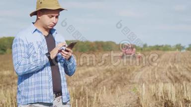 英俊的农民与智能手机站在田间向日葵与联合收割机的背景。 现代概念