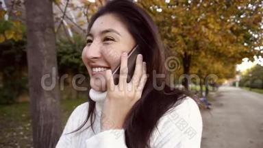 慢动作亚洲快乐女孩穿着白色毛衣，用智能手机说话，在<strong>公园里散步</strong>。