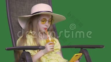 夏天喝橙汁和看智能手机的年轻女孩。 女孩坐在椅子上享受水果鸡尾酒