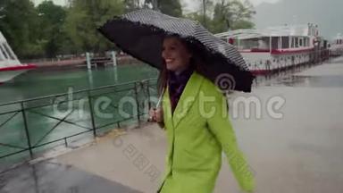 快乐的女人穿着黄色的外套，带着黑色的圆点伞，沿着白色游艇的长廊在雨中奔跑