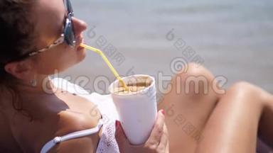 特写镜头。 肖像。 一位年轻的<strong>女士</strong>戴着<strong>太阳镜</strong>和泳衣躺在海滩上，通过一杯冷咖啡喝着