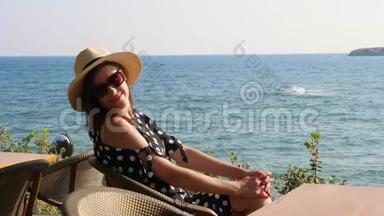 布鲁内特女孩戴着<strong>帽子</strong>和太阳镜，坐在海边的一家咖啡馆的桌子旁，欣赏海景。 炎热的<strong>夏日</strong>。