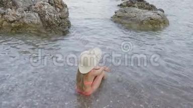 <strong>暑假</strong>，戴帽子的年轻女孩和泳装坐在鹅卵石海滩的海水中。 <strong>旅游</strong>女孩享受柔软的海浪