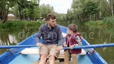 年轻的父亲和他可爱的小儿子在绿树丛中的湖上乘着蓝色的小船航行。 快乐的爸爸和儿子一起在湖边度过时光。