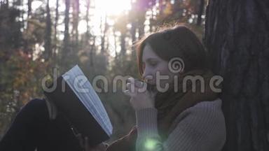 迷人的黑发女人旅行者喝热饮料从热水杯，阅读书籍，怀旧在秋天森林公园在阳光
