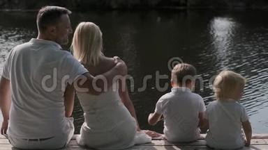 幸福的家庭<strong>爸爸妈妈</strong>和两个孩子坐在河边的码头上。 背面