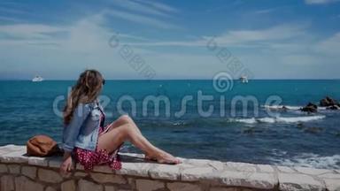 阳光明媚的一天，穿着连衣裙和牛仔夹克的年轻女子坐在石栅栏上看着<strong>蔚蓝</strong>的<strong>大海</strong>。 风的美丽发展