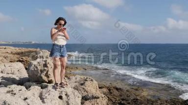 迷人的年轻女子站在海边的岩石上，看着电话。 白种人黑发女孩站在海边打电话