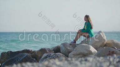 穿着绿色<strong>夹克</strong>、牛仔短裤和<strong>运动</strong>鞋的漂亮女孩坐在石滩上，享受着令人惊叹的海景。