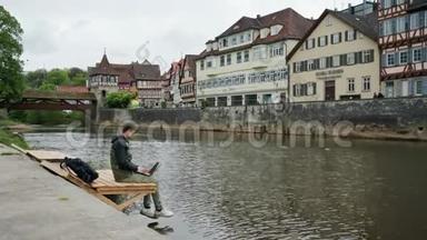 带背包的年轻人坐在靠近水的木墩上，用笔记本。 传统德语