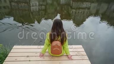长头发的女人穿着黄色夹克，背着背包坐在河边或池塘的木墩上，看上去像鸭子在游泳。 传统