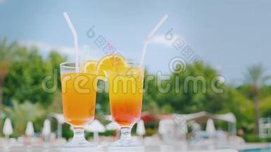 <strong>泳池</strong>边的<strong>沙滩</strong>桌上放着两杯带有吸管和橘子片的橙色鸡尾酒，映衬着蓝天