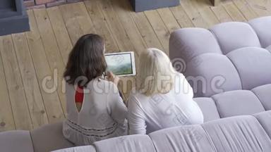 年轻<strong>女子</strong>和母亲<strong>坐在沙发上</strong>，在平板电脑上展示她的旅行照片。 友好家庭