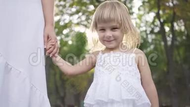 一个穿着白色裙子的小女孩站在公园或森林里，牵着成年女人的手，微笑着。 儿童保育