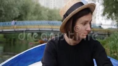 年轻的黑发女孩戴着草帽在公园里的柳树间的木船上划船。 浪漫的快乐女孩在洛杉矶划船
