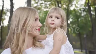 年轻的金发女人穿着白色连衣裙抚摸她可爱的女儿的肖像。 户外休闲。 家庭幸福。 一名家长