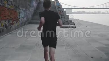 职业运动员在码头跑步的后视图。 活泼苗条的男人沿着河跑。 运动健康的小男孩在长廊上奔跑