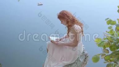 侧面看一个年轻迷人的女孩，留着长长的红色头发，在蓝湖的背景下阅读诗歌。 可爱的白种人
