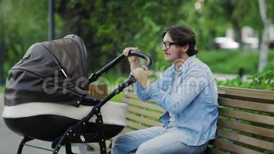 快乐的年轻爸爸欣赏刚出生的摇摆婴儿车，坐在公园的长凳上