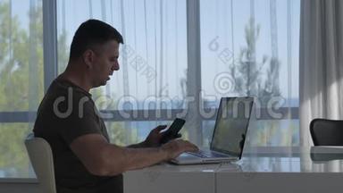 一个男人坐在酒店房间的一张桌子前，在一扇俯瞰大海的大窗户前，拿着笔记本电脑工作，在上面说话。