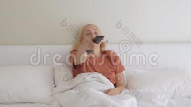 这位女士躺在家里的床上，用电子设备切换遥控电视频道，测量她的体温