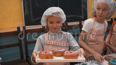 快乐女孩戴着厨师帽和围裙，在烹饪大师班上展示带有釉面甜甜圈的托盘。 带烤的可爱厨师女孩