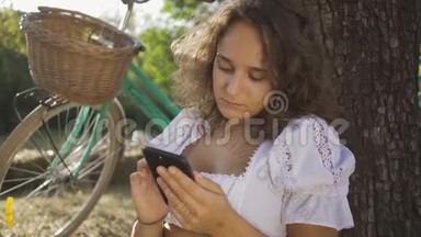 一位美丽的年轻女子，卷发坐在树下，她的手机在花园或公园里，她的自行车