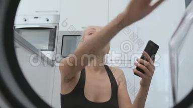 一位坐在洗衣机前的女士正在把脏衣服装进洗衣机，通过智能手机显示它是活的