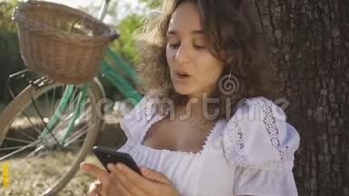 肖像可爱的年轻女子卷发坐在树下，她的手机在花园或公园里，她的自行车