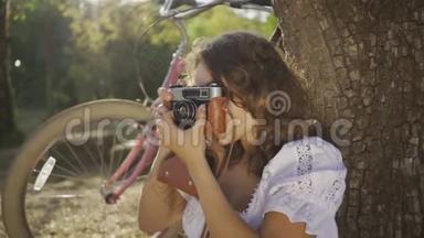 漂亮的年轻摄影师，卷发<strong>坐在树下</strong>，在花园或公园里用旧相机拍照