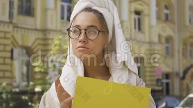 一幅可爱的年轻女人穿着浴袍，头上戴着毛巾，手里拿着文件夹，站在街上。 这就是
