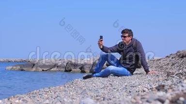 男人旅行者坐在海石海滩上，在度假时<strong>制作</strong>自拍智能<strong>手机</strong>。