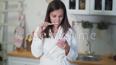 穿着<strong>白色浴袍</strong>的年轻女子在早晨刷牙，使用智能手机