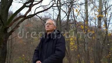 一个头发花白、戴着眼镜的瘦长男子站在森林里，在秋天的森林里挽起外套取暖