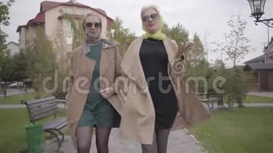 两个穿着米色外套、<strong>短裙</strong>和太阳镜的成熟白种人妇女在城市公园散步和交谈。 魅力