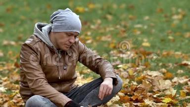 迷人的家伙坐在公园里的落叶上，仔细地拿起一片黄色的枫叶，仔细地检查它。