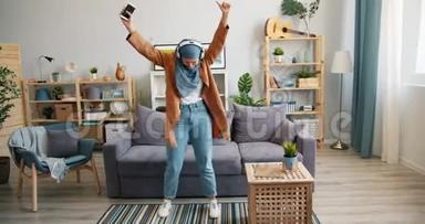 快乐的中东女人戴着头巾在家跳舞带着耳机智能手机