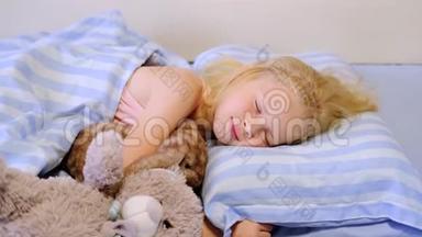 一个漂亮的金发女孩穿着粉红色睡衣，一大早就睡在床上，床上铺着蓝色的床上用品。 女孩和她的泰迪熊