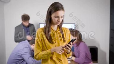 年轻女子站在办公室的智能手机上打字，同事们坐在后台