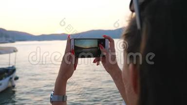 女人在他的智能手机上用金色日落拍摄风景优美的海洋景观。 女孩用手机变漂亮