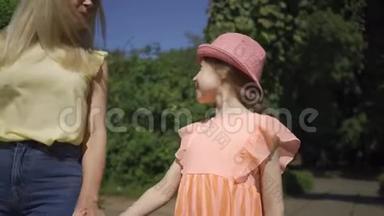 可爱的金发母亲和可爱的小女儿在夏天的花园里牵手散步。 家庭幸福。 妇女和女孩