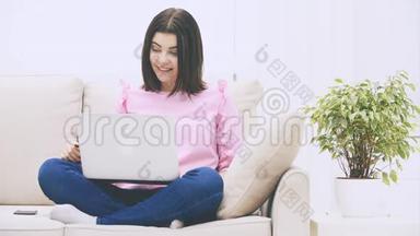 可爱的白种人黑发女孩坐在沙发上，拿着笔记本电脑，在互联网上冲浪，气