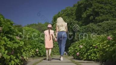 后景可爱的金发妈妈和小可爱的女儿在夏天的花园里牵手散步。 幸福家庭