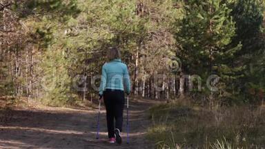 女人练习北欧在秋天的森林里行走，她`在沙地路上远离相机。 后景