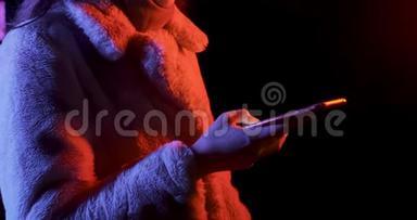 漂亮的黑发女人穿着红色和蓝色霓虹灯在晚上看智能手机。 时尚模特女孩化妆和