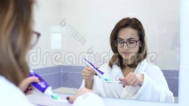 一个戴眼镜的年轻女人刷牙。 一个穿着白色外套的女人开始刷牙。 从镜子里看到